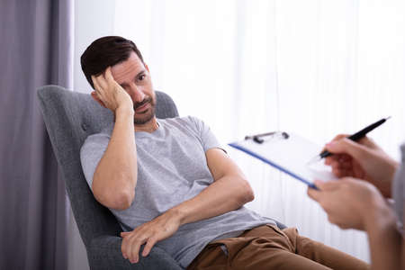 Female Psychologist With Clipboard Sitting Near Young Man Suffering From Depression