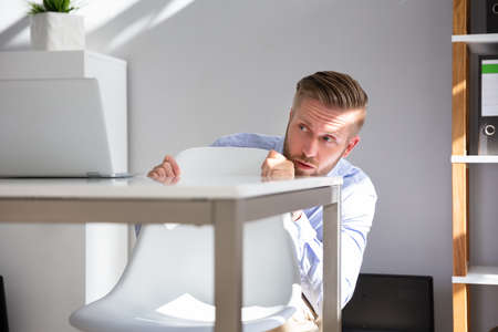 Afraid Businessman Peeking From The Edge Of Desk In Office