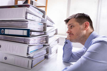 Rear View Of Stressed Businessman With Hand On Head In Front Of Stacked Folders