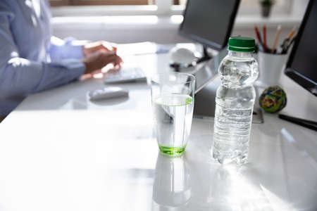 Water Bottle And Drinking Glass On Desk And Man In Background Using Computer