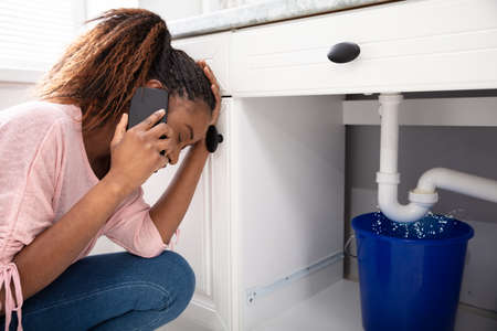 Young Woman Calling Plumber Crouching In Front Of Water Leaking From Sink Pipeの写真素材