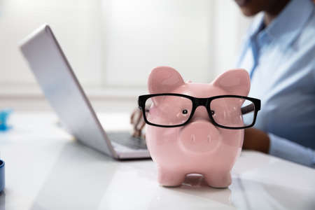 Close-up Of Pink Piggy Bank In Front Of A Business Person Using Laptop At Workplaceの写真素材