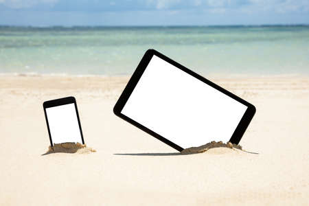 Mobile Phone And Digital Tablet Dig On Sandy Beach Near The Idyllic Seaの写真素材