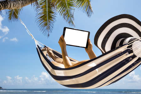 Close-up Of A Woman Wearing Hat Lying On Striped Hammock Using Digital Tablet At Beachの写真素材