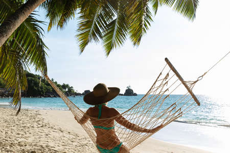 Rear View Of Young Woman In Bikini And Hat Sitting On Hammock On The Beachの写真素材