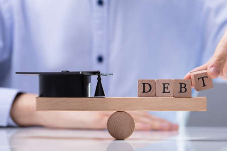Businessperson's Hand Putting Word Debt On Wooden Seesaw To Balancing Black Graduation Hatの写真素材