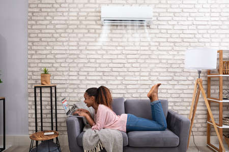 Happy Young African Woman Using Laptop Enjoying Cooling Under Air Conditioner At Homeの写真素材