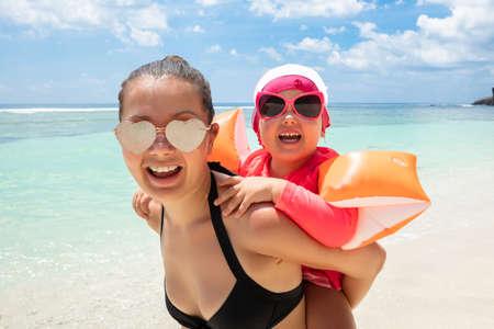 Portrait Of A Daughter Enjoying The Piggyback Ride On Her Mother's Back At Beach