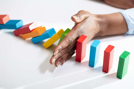 Close-up Of A Businesswoman Hand Stopping Colorful Dominoes From Falling On Office Deskの写真素材