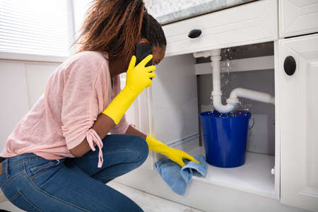 Close-up Of A Young Woman Placing Blue Bucket Under Water Leaking From Sink Pipeの写真素材