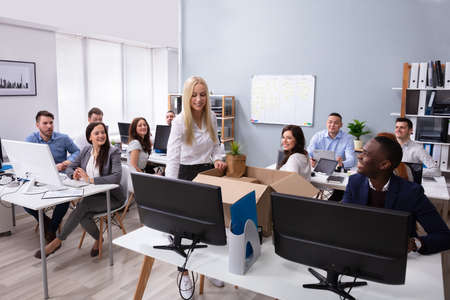 Businesspeople Looking At Smiling New Employee Unpacking Box With Personal Belongings In Office
