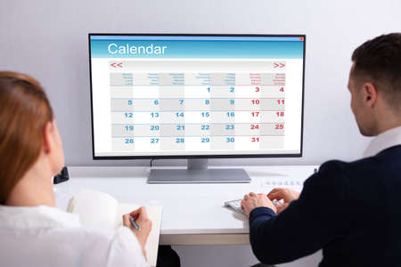 Two Businesspeople Looking At Calendar On Desktop Computer At Desk