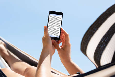 Woman Relaxing On Hammock Reading The Book On Mobile Phone Against Blue Sky