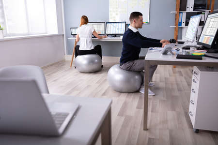 Two Businesspeople Working On Computer In Office Sitting On Fitness Ball