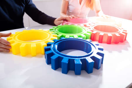 Two Businesspeople Joining Colorful Gears Over White Desk In Officeの写真素材