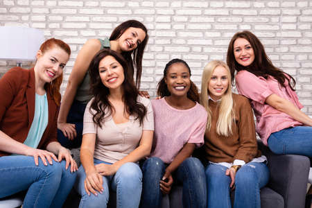 Portrait Of Beautiful Smiling Women Sitting On Sofa Against Brick Wallの写真素材