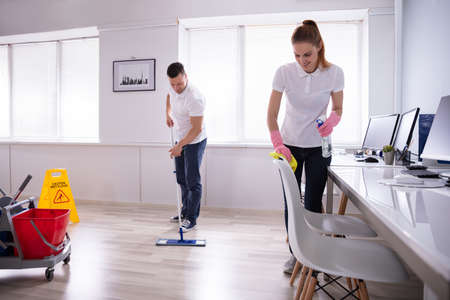 Two Smiling Young Janitor Cleaning The Desk And Mopping Floor In The Officeの写真素材
