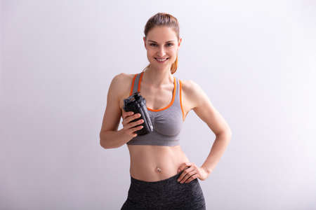 Portrait Of Smiling Fitness Woman Holding Protein Shaker Bottle Against Gray Backgroundの写真素材