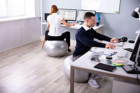 Two Businesspeople Working On Computer In Office Sitting On Fitness Ballの写真素材