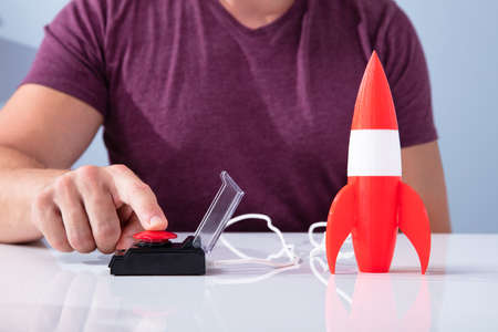 Man's Hand Launching Rocket By Pressing Red Button Over Reflective Desk