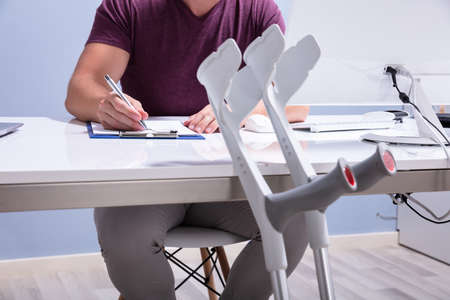 Close-up Of Crutches Leaning On Table In Front Of Handicapped Man Filling Form