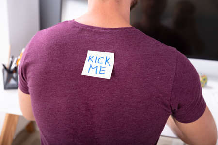 Rear View Of Man Working In Office With Kick Me Text Written On Sticky Note