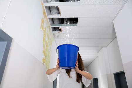 Worried Woman Holding A Blue Bucket Under The Leak Ceiling In Corridor