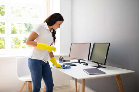 Side View Of Female Janitor Cleaning Desk With Blue Napkin In Officeの写真素材
