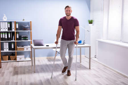 Handicapped Businessman Walking On Hardwood Floor With Crutches At Workplaceの写真素材