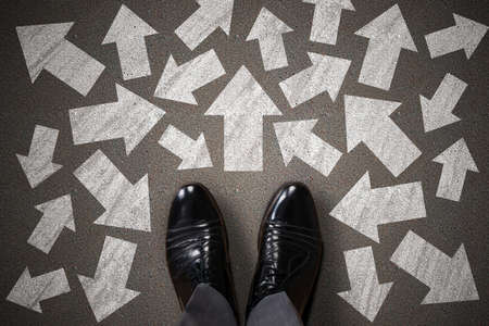 High Angle View Of Businessperson Standing On Asphalt With Different Directional White Arrow Signs