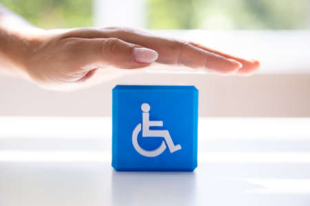 Close-up Of A Person's Hand Protecting Blue Cubic Block With Disabled Icon