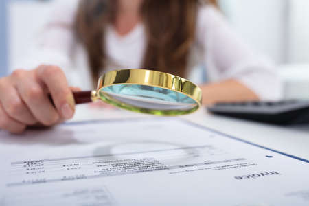 Close-up Of A Businesswoman Looking Invoice Through Magnifying Glass On Desk In Officeの写真素材