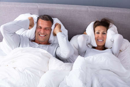 Mature Couple Disturbed By Noise Covering Their Ears With Pillow On Bed