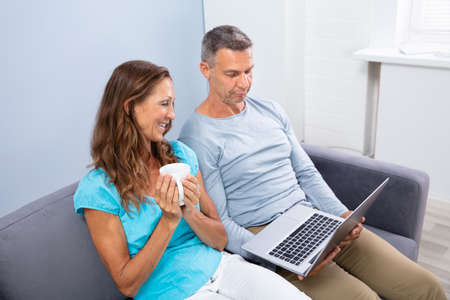 Happy Mature Couple Sitting On Sofa Watching Movie In Laptop At Home