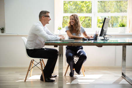 Smiling Male And Female Colleagues Sitting At Office Desk Working Together While Using Computerの写真素材