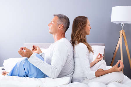 Matured Couple Sitting Back To Back On Bed Meditatingの写真素材