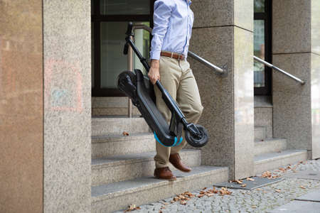 Man Carrying E-Scooter From Work Walking Out Of Office Building
