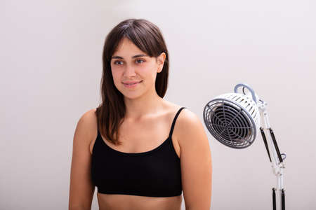 Smiling Young Woman Receiving Infrared Radiation Heat Light Lamp Therapy Against Gray Background