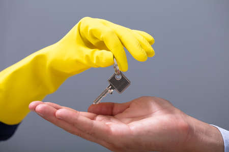 Man's Hand Receives Keys From Cleaner's Hand Wearing Yellow Gloves Against Gray Backgroundの写真素材