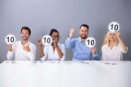 Panel Of Excited Judges Sitting In Front Of Grey Wall Showing 10 Score Cards