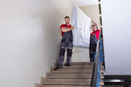 Two Young Male Movers In Uniform Carrying The Wrapped Mattress While Moving Downward The Staircaseの写真素材