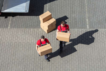An Elevated View Of Two Young Male Movers Walking With Cardboard Boxes On Cobblestone Pavement