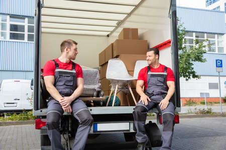 Portrait Of Smiling Movers Sitting In Van With Stack Of Cardboard Boxes And Furniture