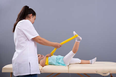 Girl Stretching The Leg With Yellow Elastic Band Instructed By A Female Physiotherapist In Clinicの写真素材