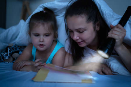 A Mother Tells Stories To Daughter Under The Blanket With A Illuminated Torch On Bedの写真素材