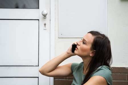 An Afraid Young Woman Sitting Outside The Door Talking On Mobilephoneの写真素材