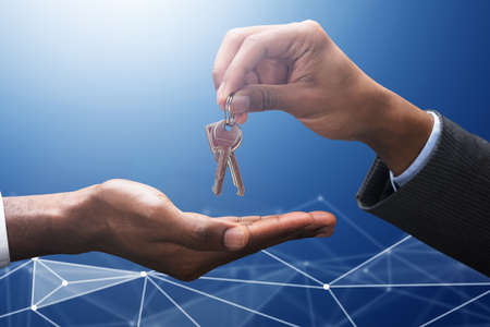 Close-up Of A Businessman Giving Keys To Other Male Person's Hand Against Blue Technology Background