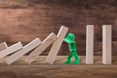 Side View Of A Green Plastic Figure Stopping The Wooden Domino Effect On Plankの写真素材
