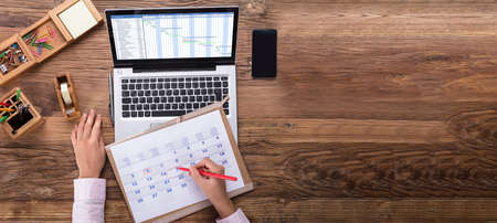 An Overhead View Of Businesswoman Marking Date On Calendar With Gantt Chart On Laptop Screenの写真素材