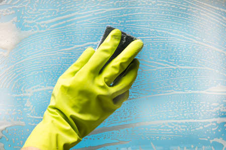 Person Wearing Green Gloves Cleaning Soap Suds Over Glass Window With Sponge